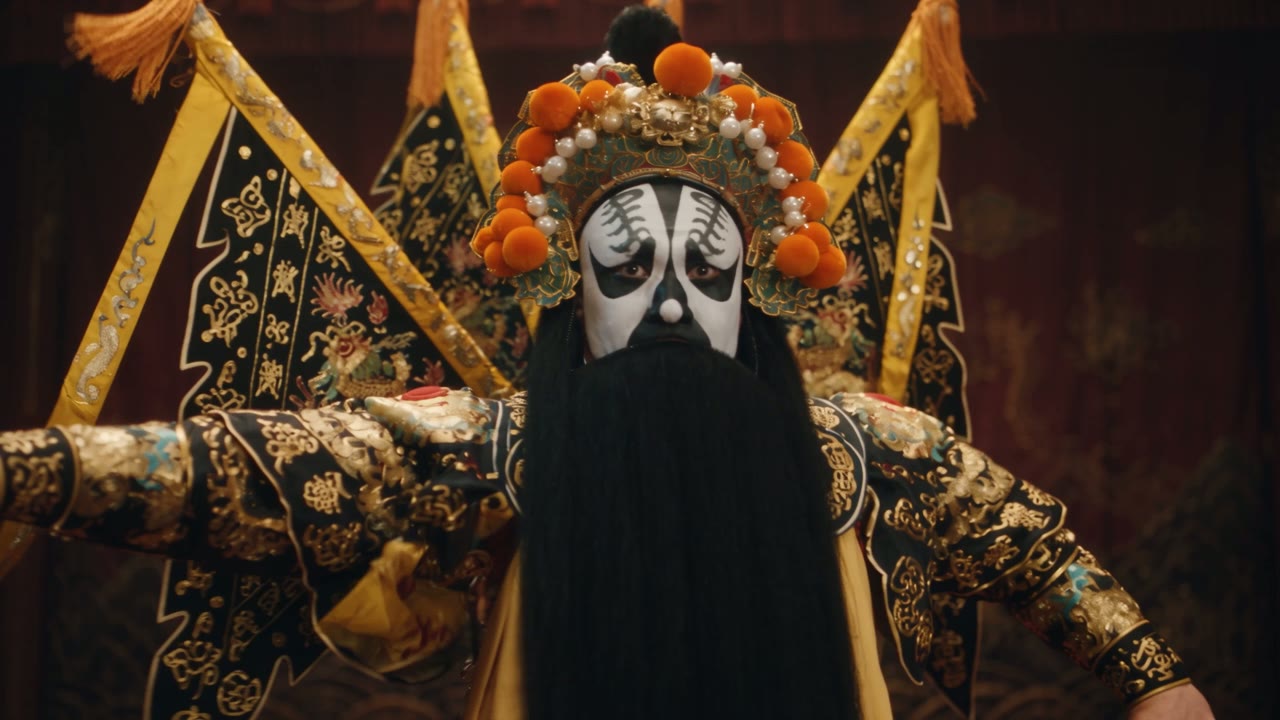 Frame from an AI video model comparison clip: a Peking opera performer in a painted mask and gold embroidered costume on a dark stage.