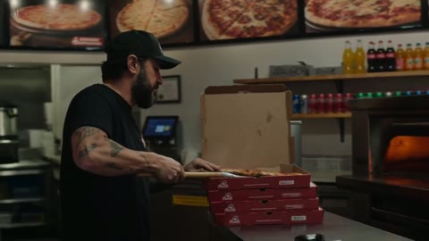 Reference image: a pizza maker in a black cap and T-shirt boxing a pizza behind the counter.