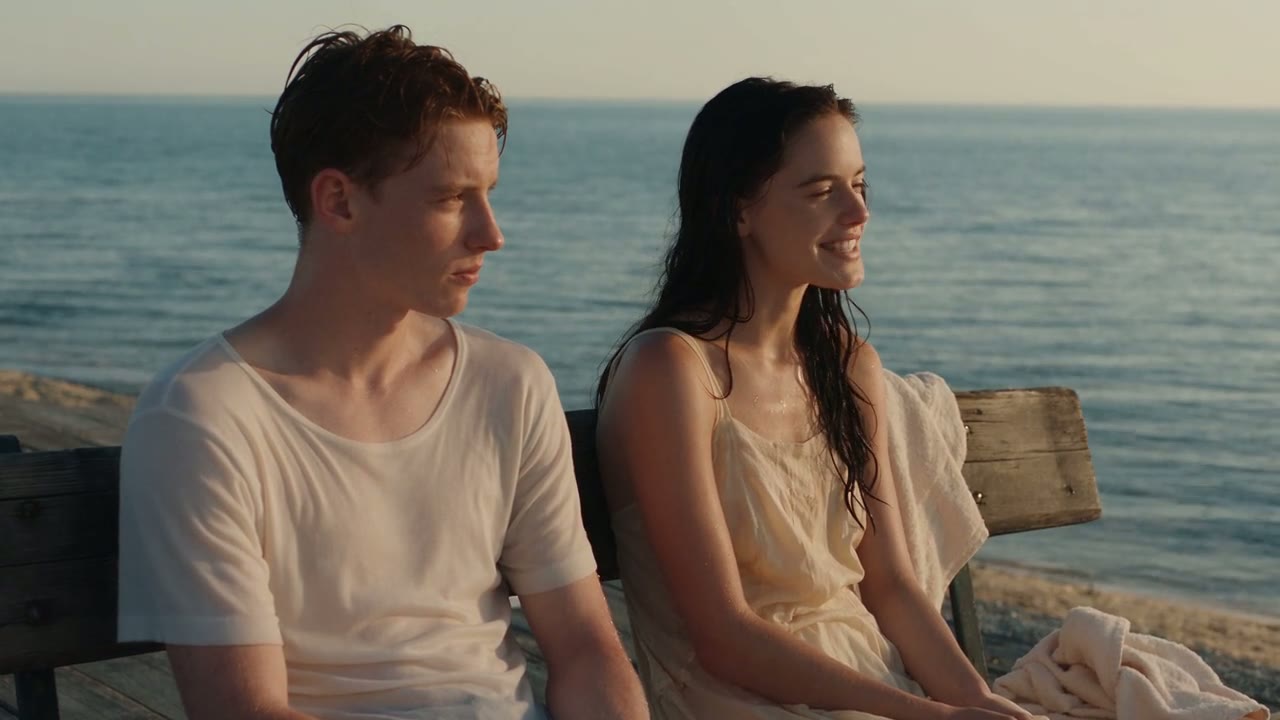Frame from a Seedance 2.5 showcase clip: two young adults sitting on a seaside bench at dusk after a swim.